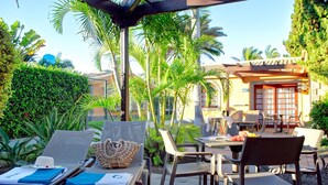 A patio area with lounge chairs, a table with chairs, and a building with a tiled roof. - Suites & Villas by Dunas (San Bartolomé de Tirajana)
