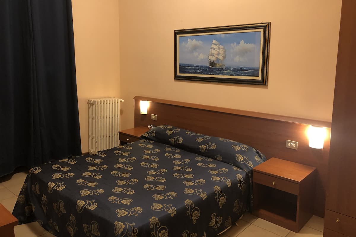 A hotel room with a bed, two bedside tables, a nightstand, a framed picture of a ship on the wall, and a radiator.