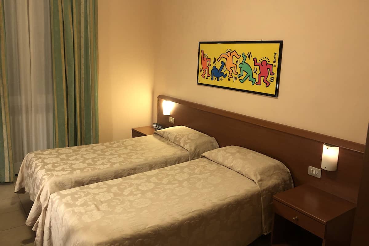A hotel room with two beds, a nightstand with a phone, a wall-mounted light, and a colorful artwork.