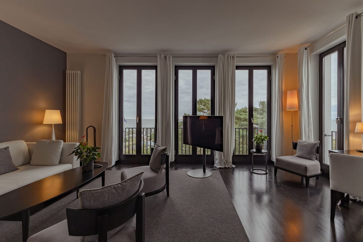 A modern hotel room with a sofa, chairs, a TV, and large windows offering a view of the outdoors.
