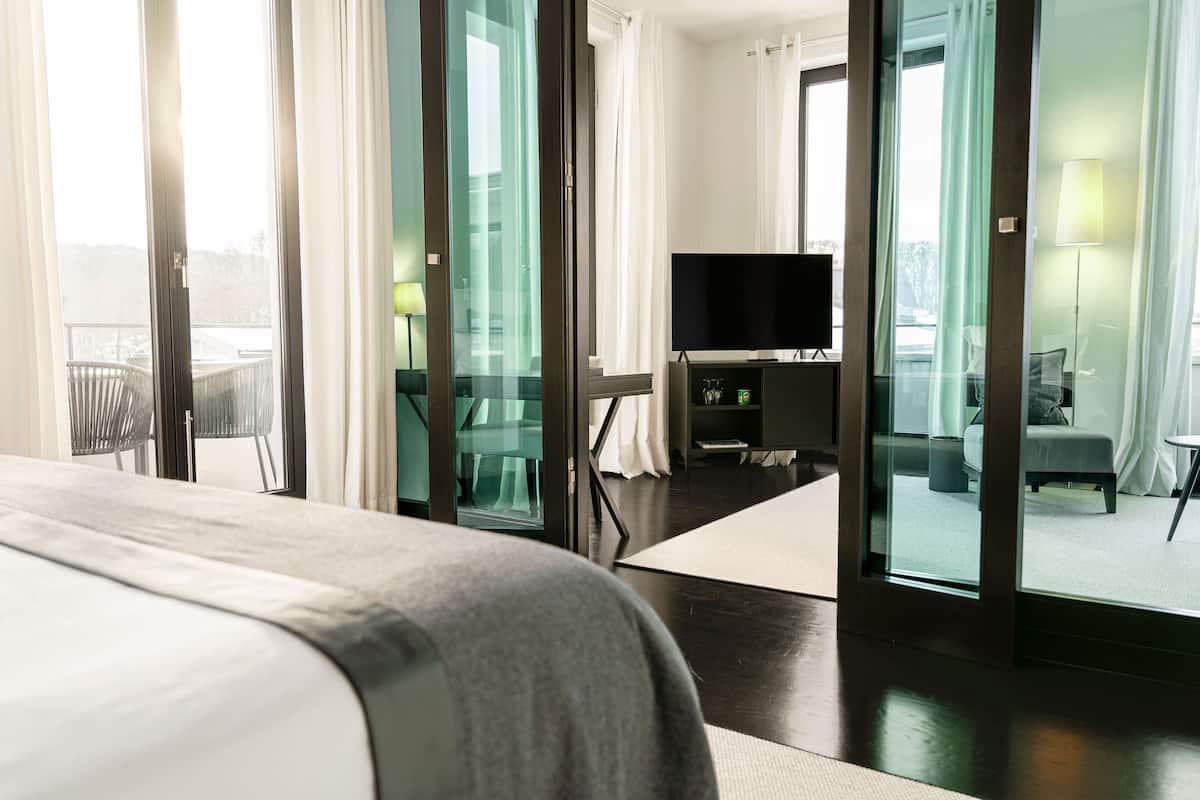 A modern hotel room with a large bed, a flat-screen TV, a desk, and a balcony with a glass door.