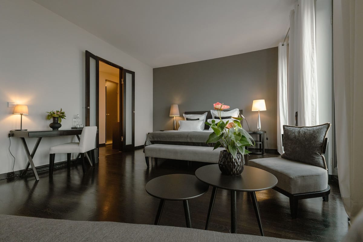 A modern hotel room with a bed, a desk, two chairs, a small table, and a potted plant.