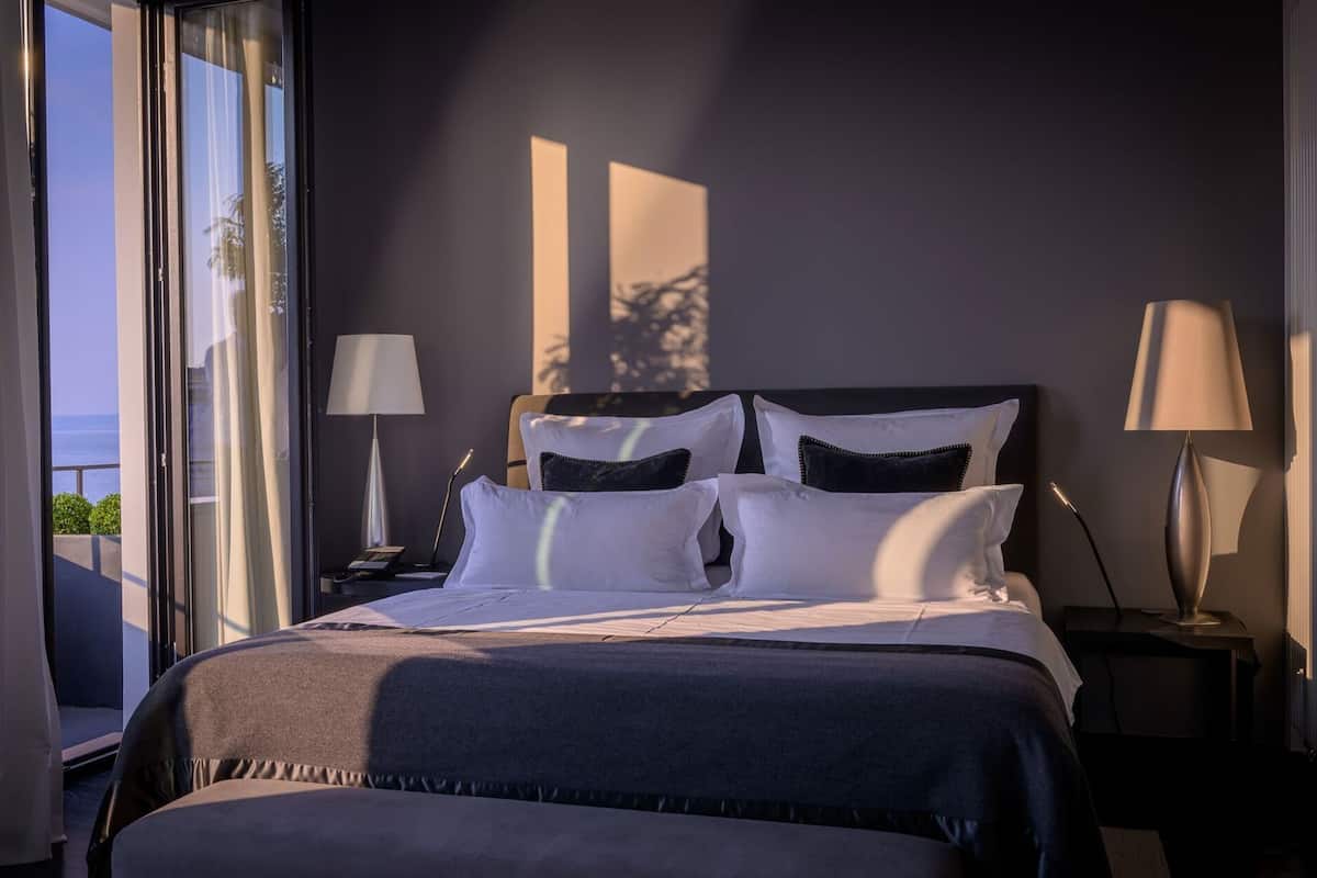 A hotel room with a bed, two bedside lamps, a nightstand, and a view of the sea.