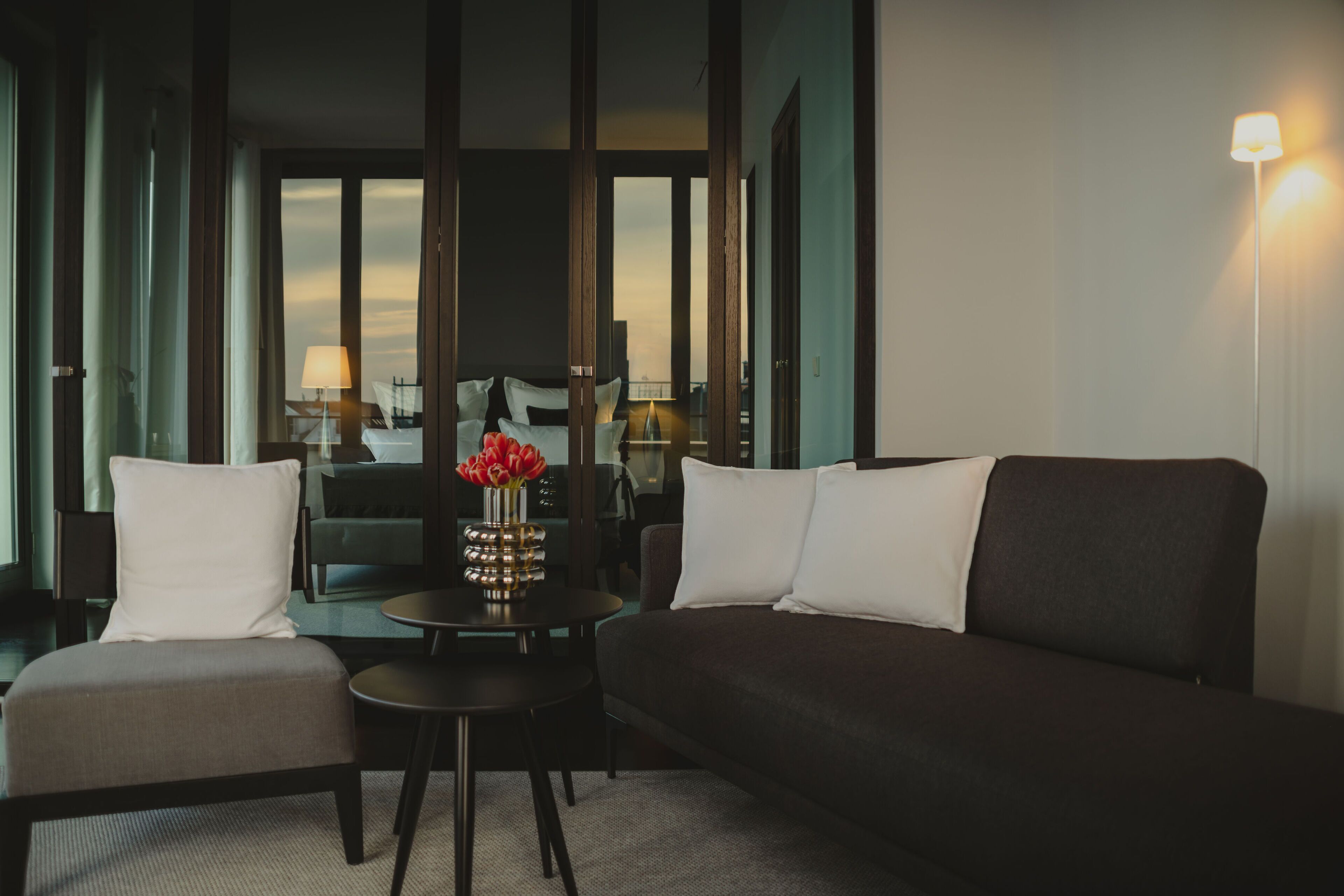 Une chambre d’hôtel moderne avec un canapé de couleur sombre, une petite table ronde et une chaise. Un vase rempli de fleurs rouges est posé sur la table.