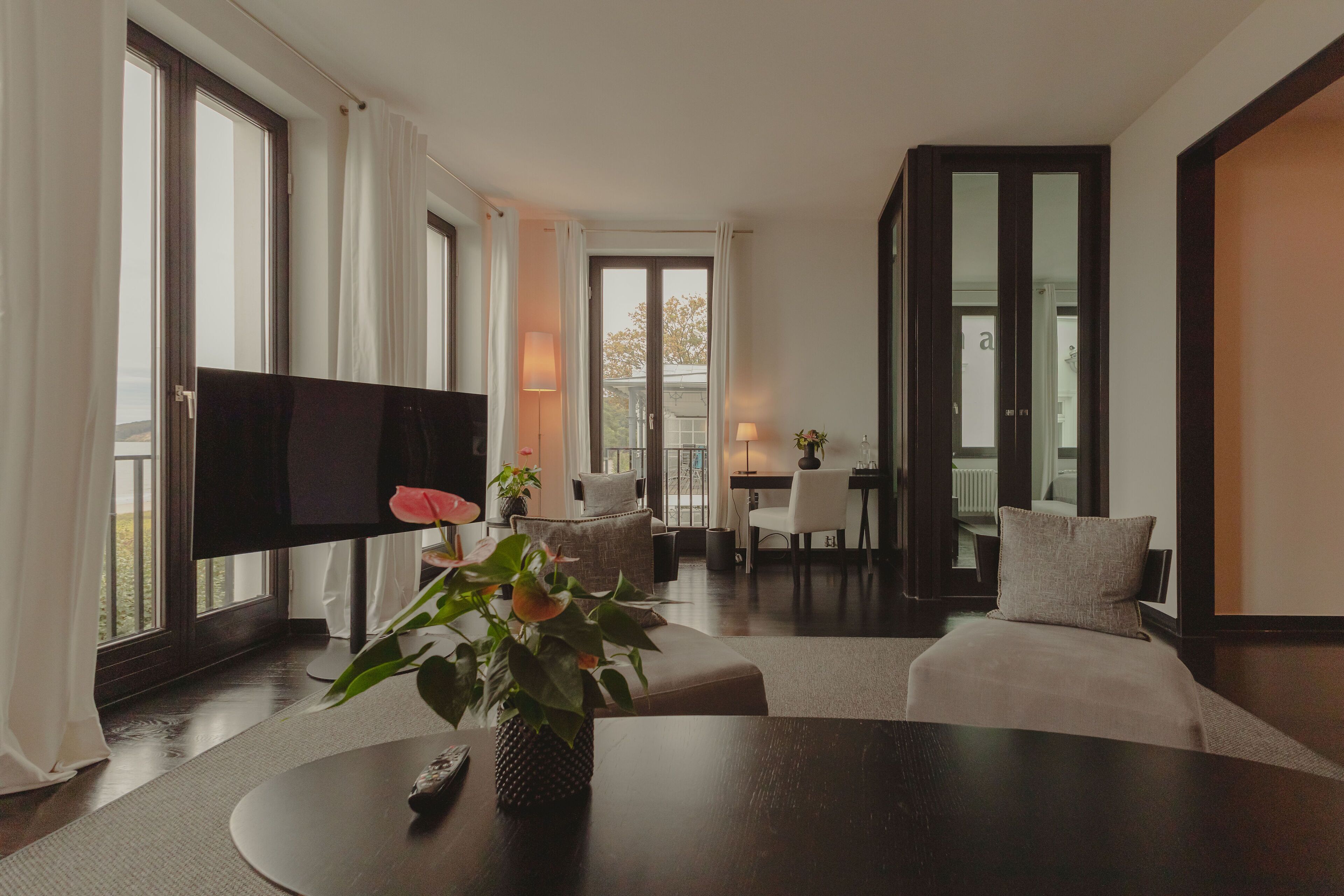 A modern living room with a flat-screen TV, a round coffee table with a vase of flowers, a sofa, and a dining area with a table and chairs.