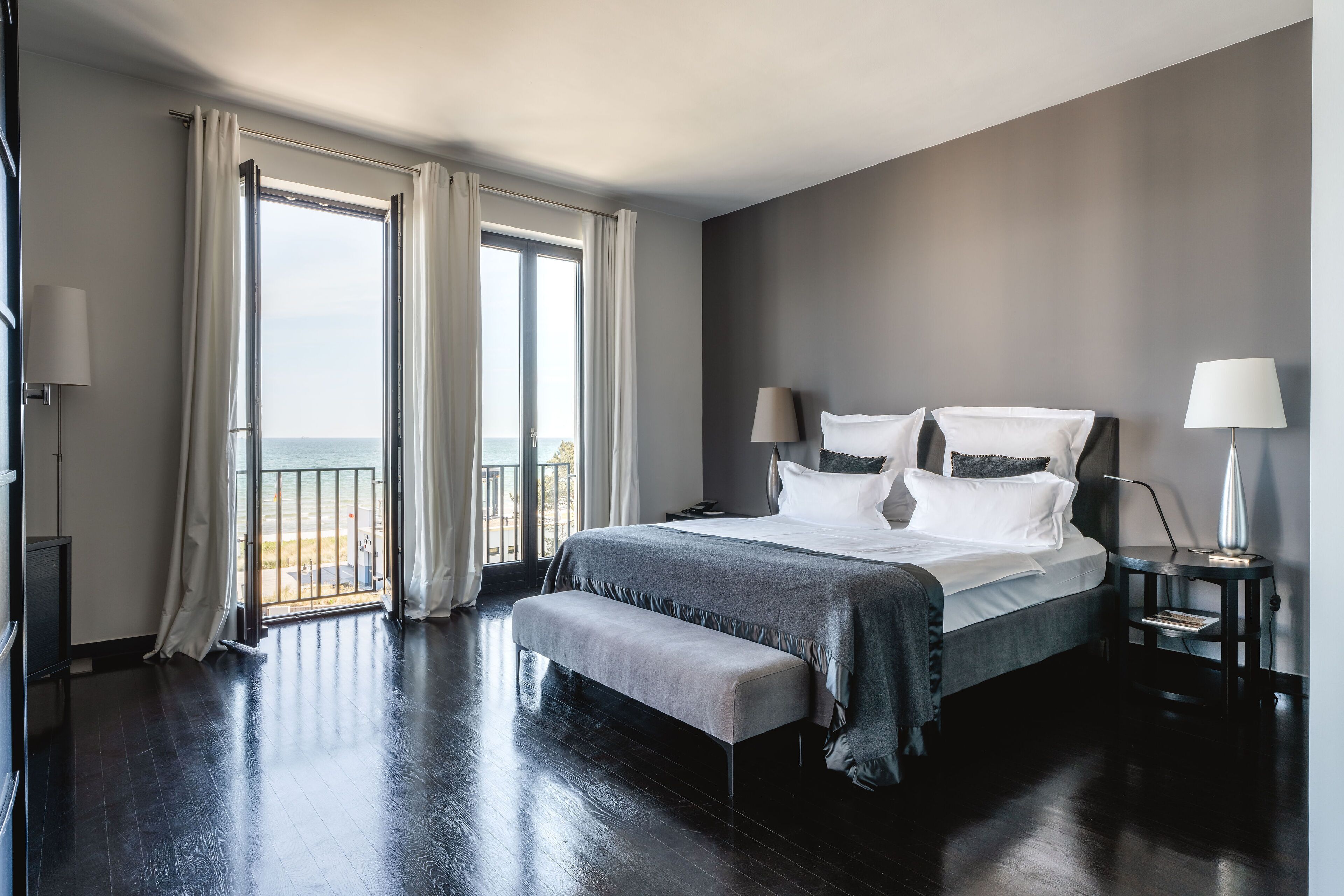 A modern hotel room with a large bed, a bench, two bedside lamps, and a view of the sea through a balcony.