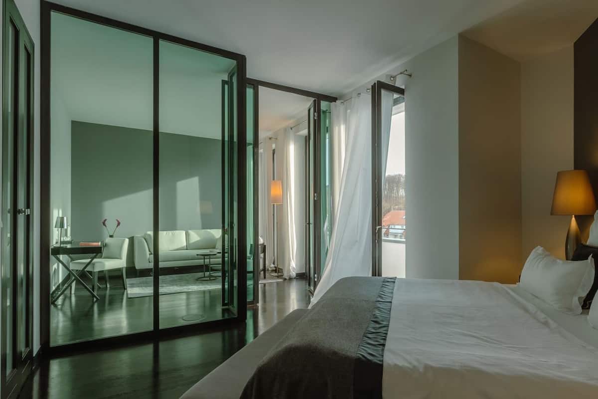 A hotel room with a bed, a desk, a chair, a sofa, and a sliding glass door leading to a balcony.
