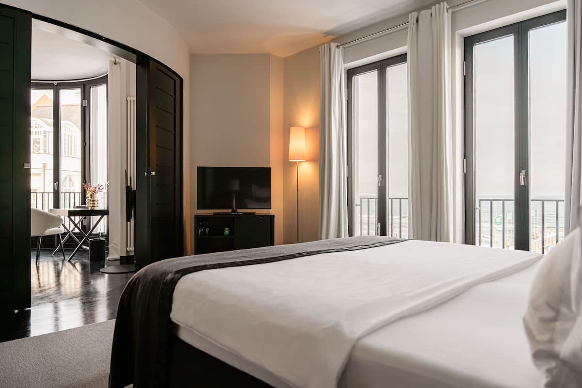 A hotel room with a large bed, a TV, a desk with a chair, and a balcony with curtains.