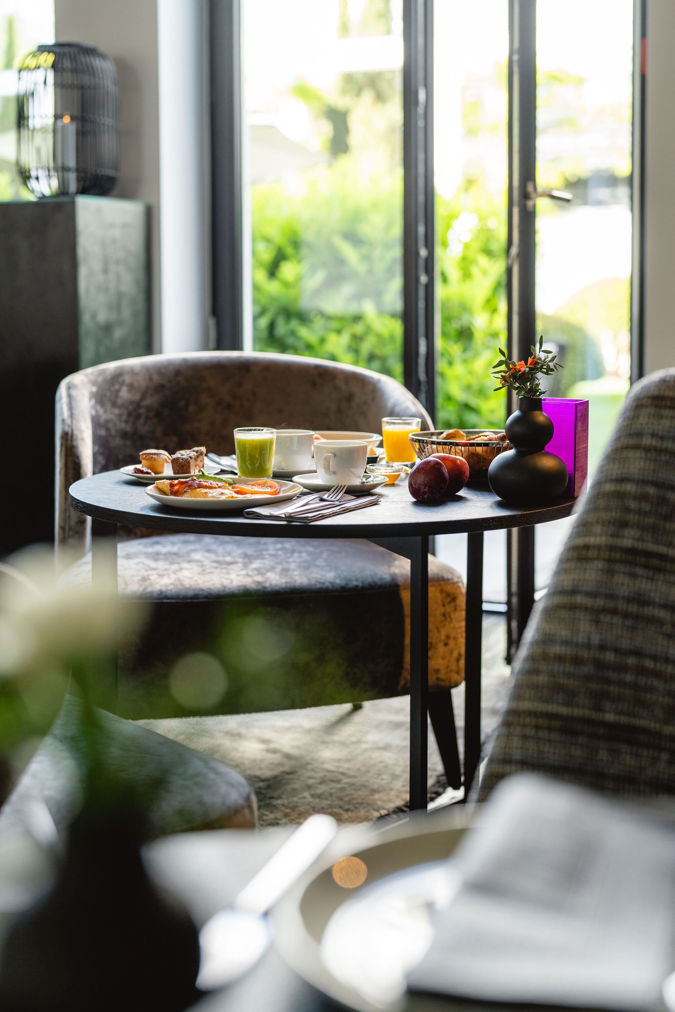 Une table de petit-déjeuner avec une variété de plats et de boissons, un vase rempli de fleurs et une vue sur la verdure à l’extérieur.