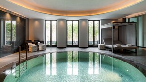Piscina interior moderna con superficie de agua cristalina, rodeada de sillas de descanso y una zona de estar con vista a zonas verdes.