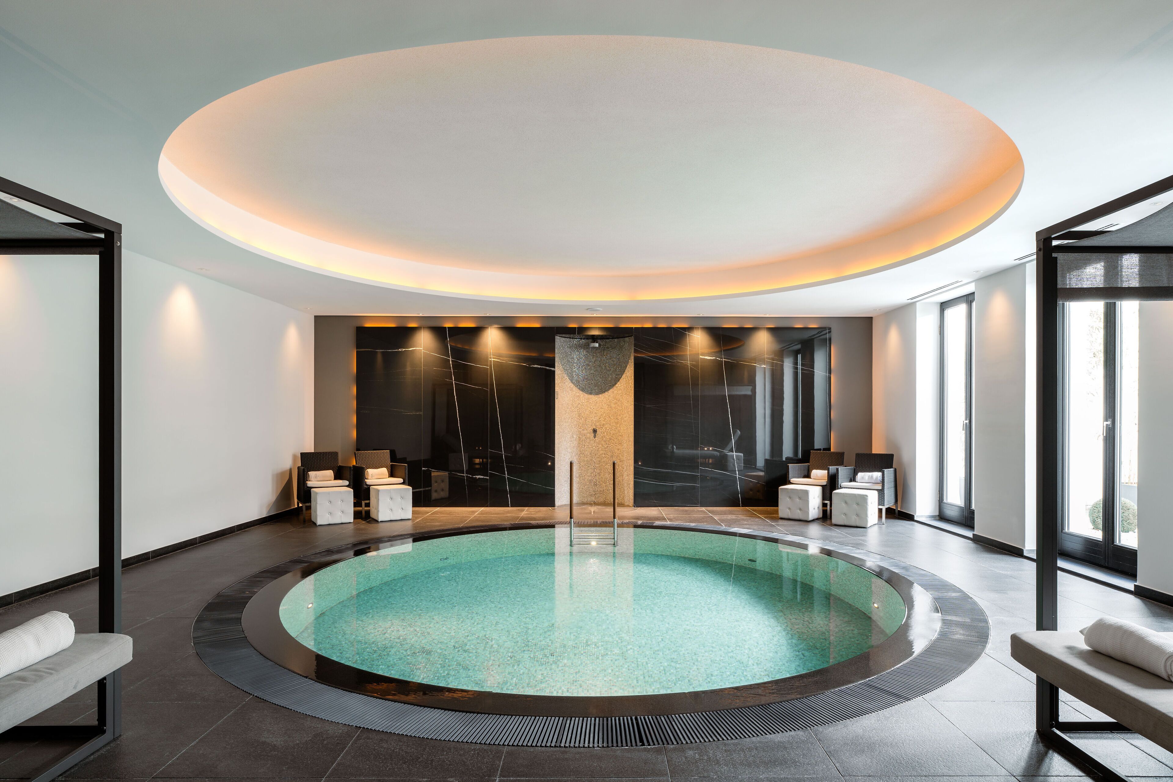 A modern indoor swimming pool with a circular design, surrounded by seating areas and a large, illuminated ceiling.