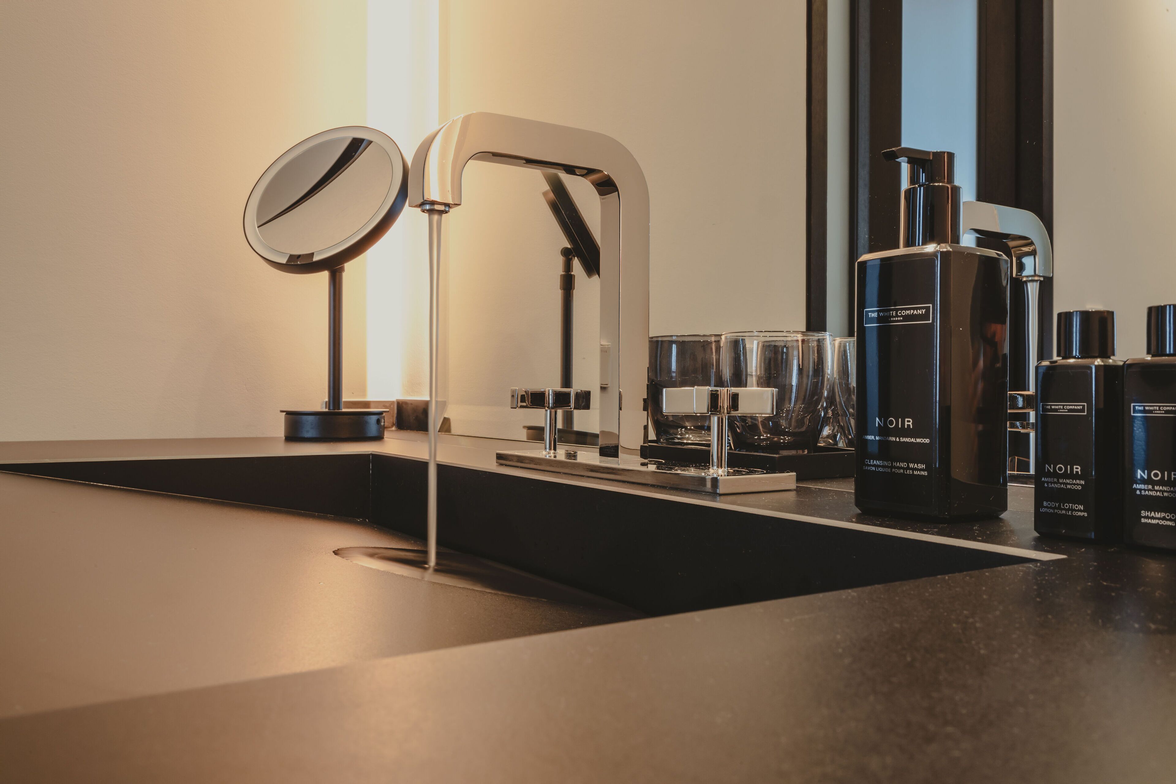 A modern bathroom with a sleek black sink, a large mirror, and a built-in faucet. There are several bottles of Noir body lotion and shampoo on the counter.