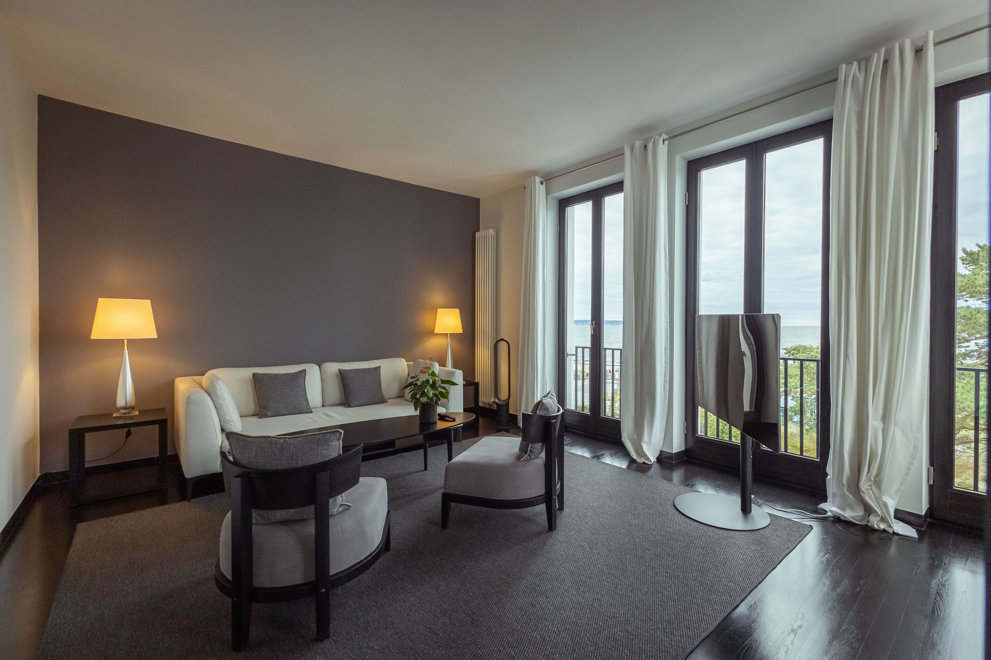Habitación de hotel moderna con sofá gris, dos sillones, una mesita y televisor. Amplias ventanas con cortinas ofrecen vistas al exterior.