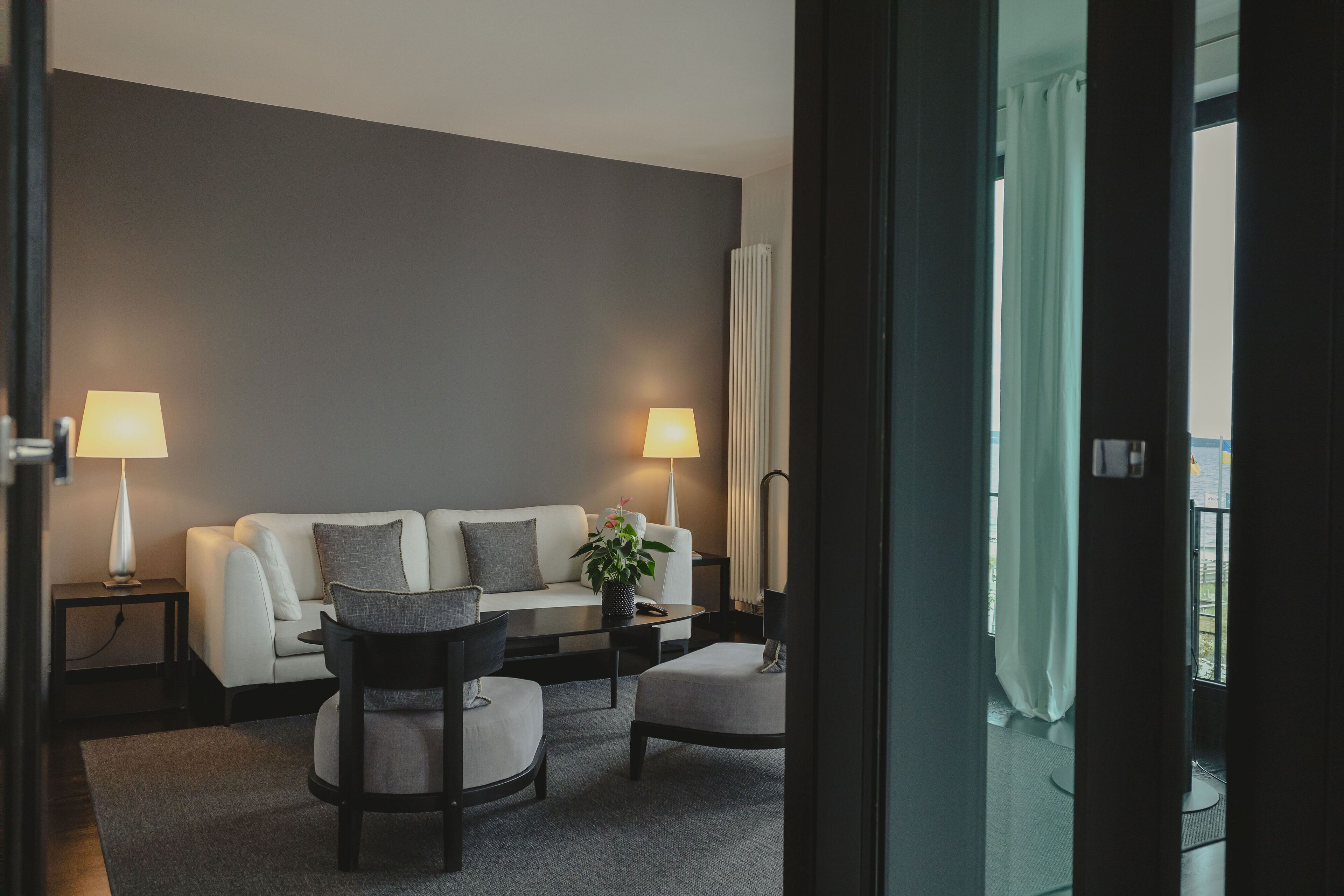 En moderne stue med sofa, to sofaborde med lamper, et sofabord og udsigt til det fri via en balkon.