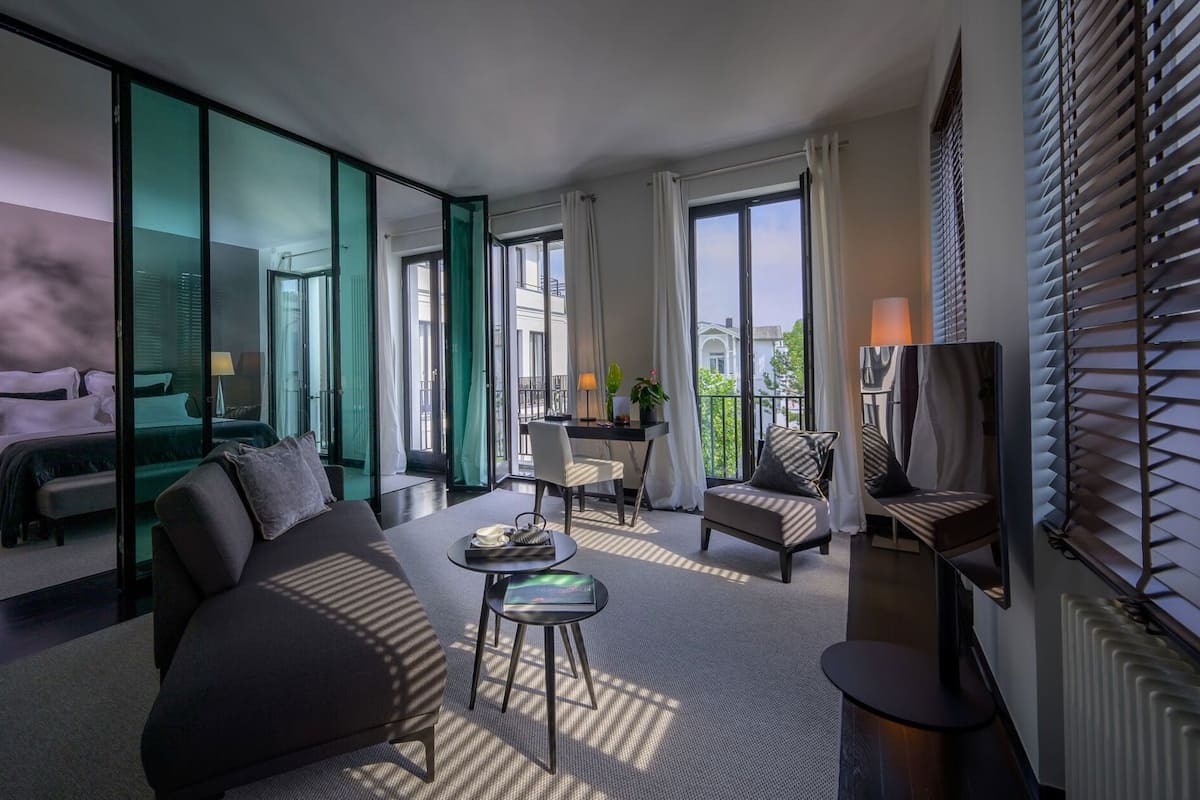 A modern hotel room with a bed, sofa, dining table, chairs, and a TV.