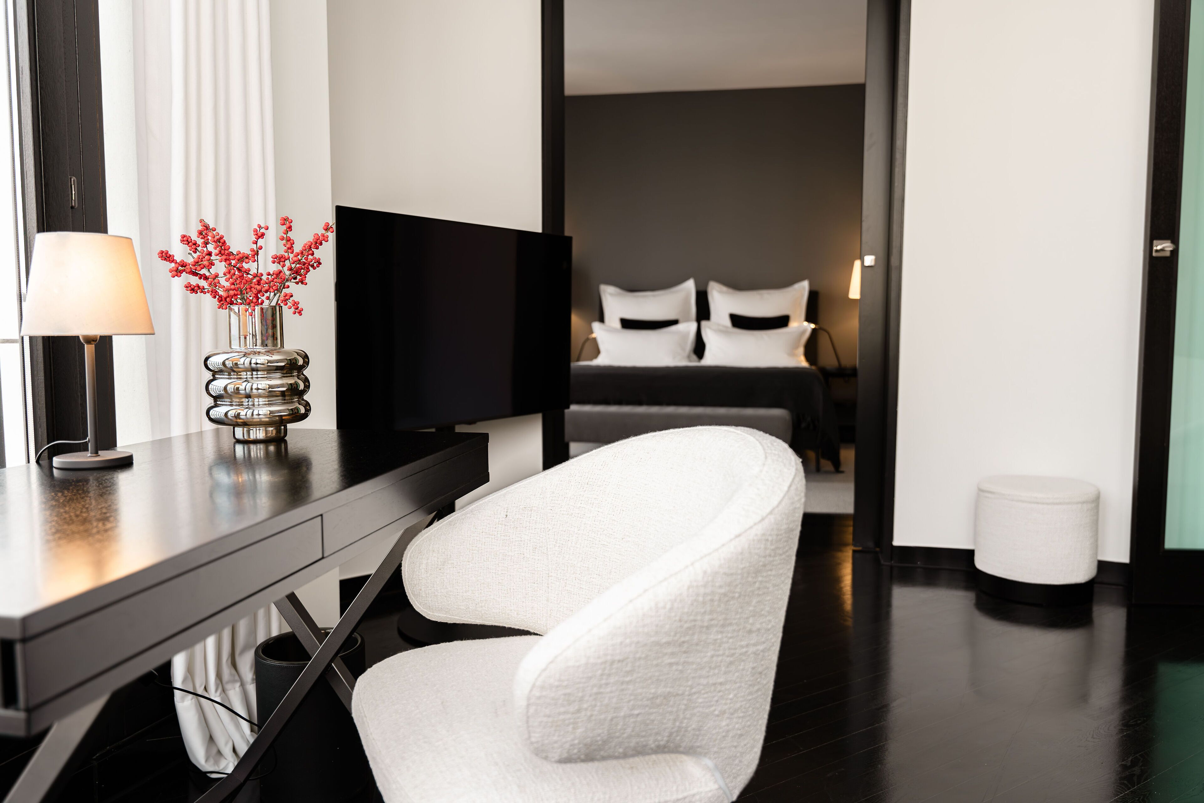 A modern hotel room with a desk, a chair, a TV, a bed with pillows, and a small white stool.