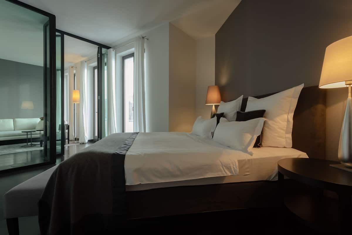 A modern hotel room with a large bed, bedside lamps, a glass door leading to a balcony, and a view of the city.
