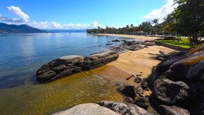 A beach with clear water, large rocks, and a distant mountain range. -  Maison Joly Residence (Ilhabela)