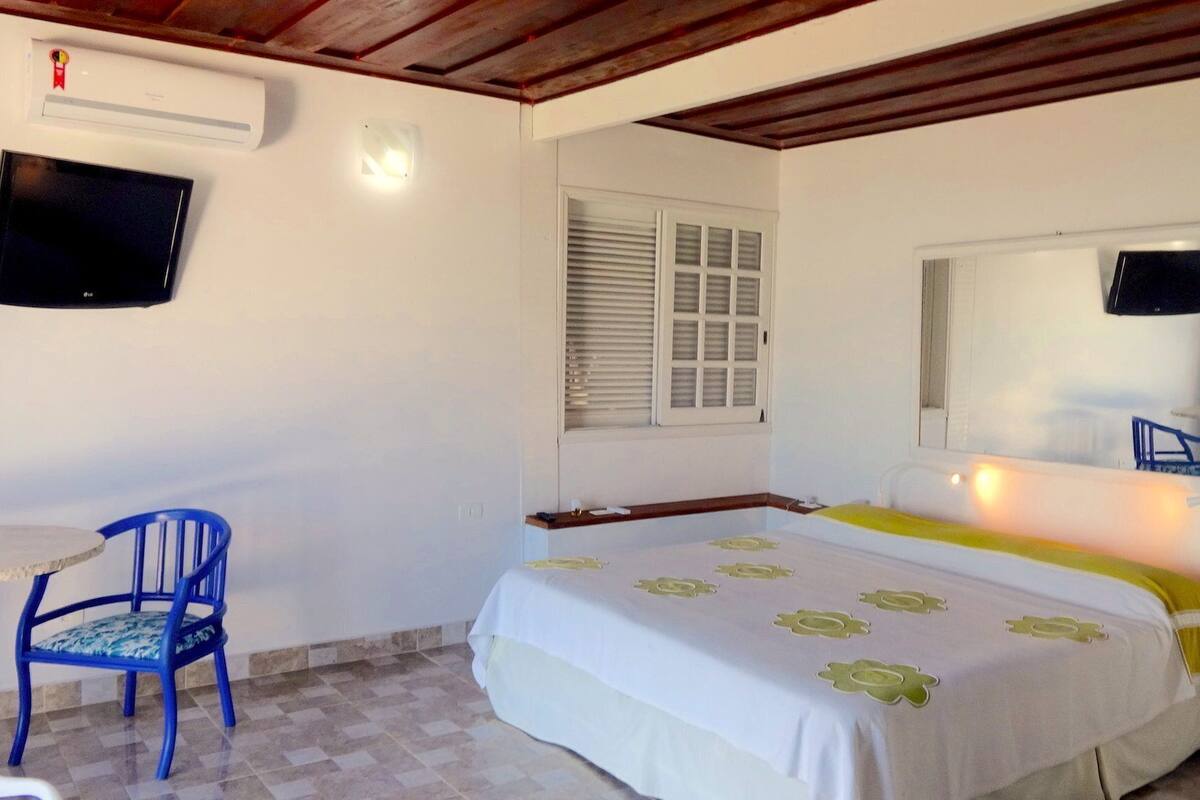 A bedroom with a bed, a chair, a window with shutters, a mounted TV, and a ceiling fan.
