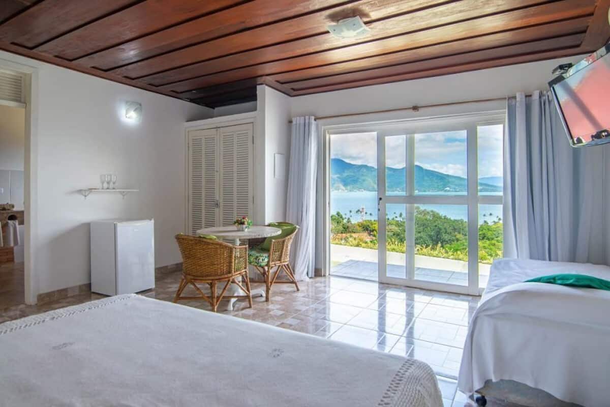 A hotel room with a bed, a dining area with a table and chairs, a refrigerator, and a sliding glass door leading to a balcony with a scenic view.