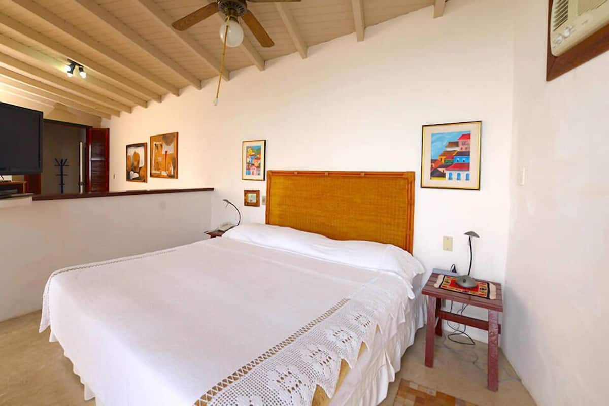 A bedroom with a large bed, a ceiling fan, and a small bedside table with a lamp and a book.