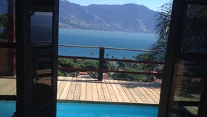 An open door leading to a wooden deck with a pool, overlooking a mountainous coastline. -  Maison Joly Residence (Ilhabela)