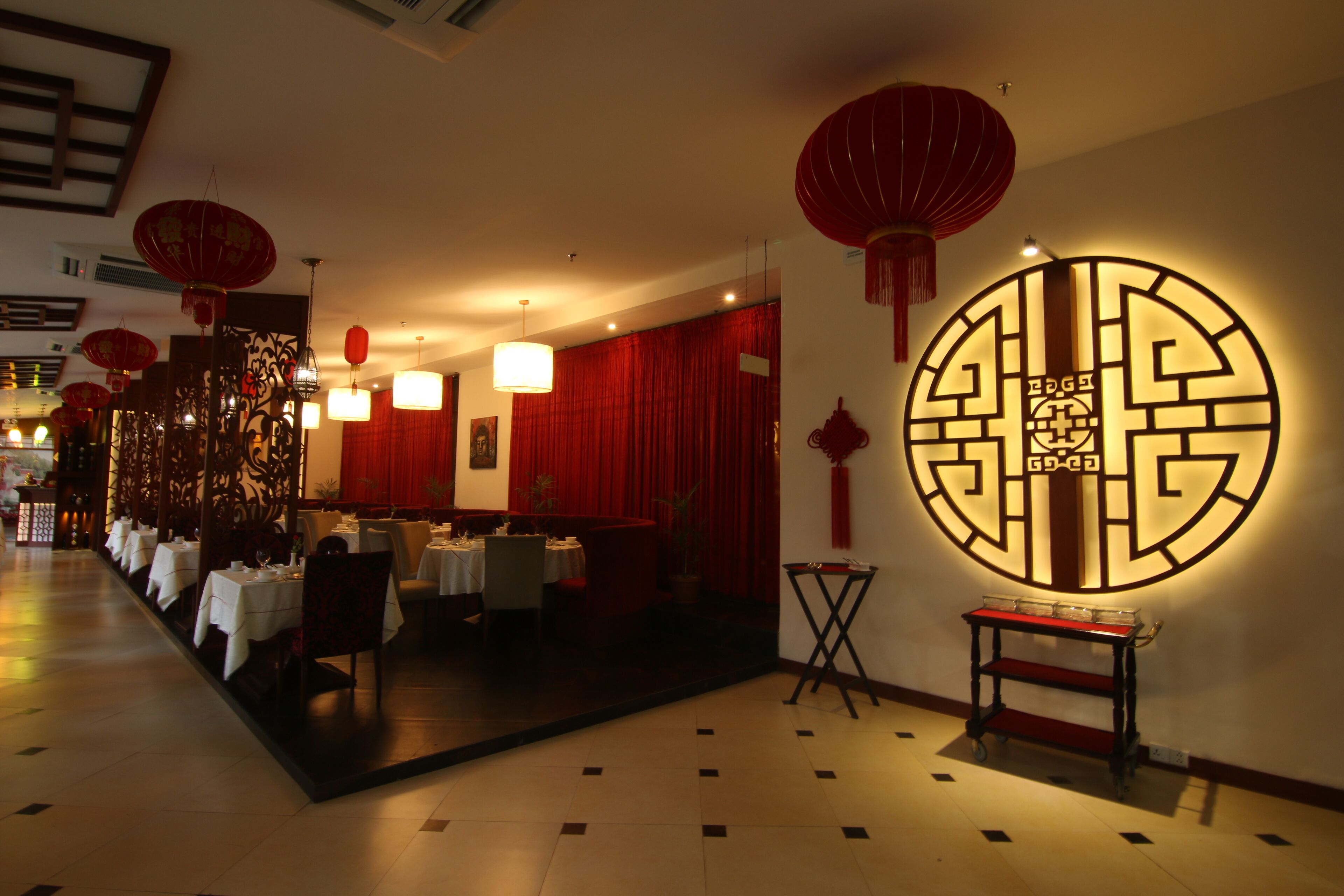 A restaurant with tables set for dining, a decorative wall with a circular emblem, and red lanterns hanging from the ceiling.
