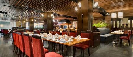 A modern restaurant with a long dining table, red upholstered chairs, and a contemporary ceiling design.