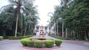 A circular fountain surrounded by neatly trimmed bushes, with palm trees and a building in the background.