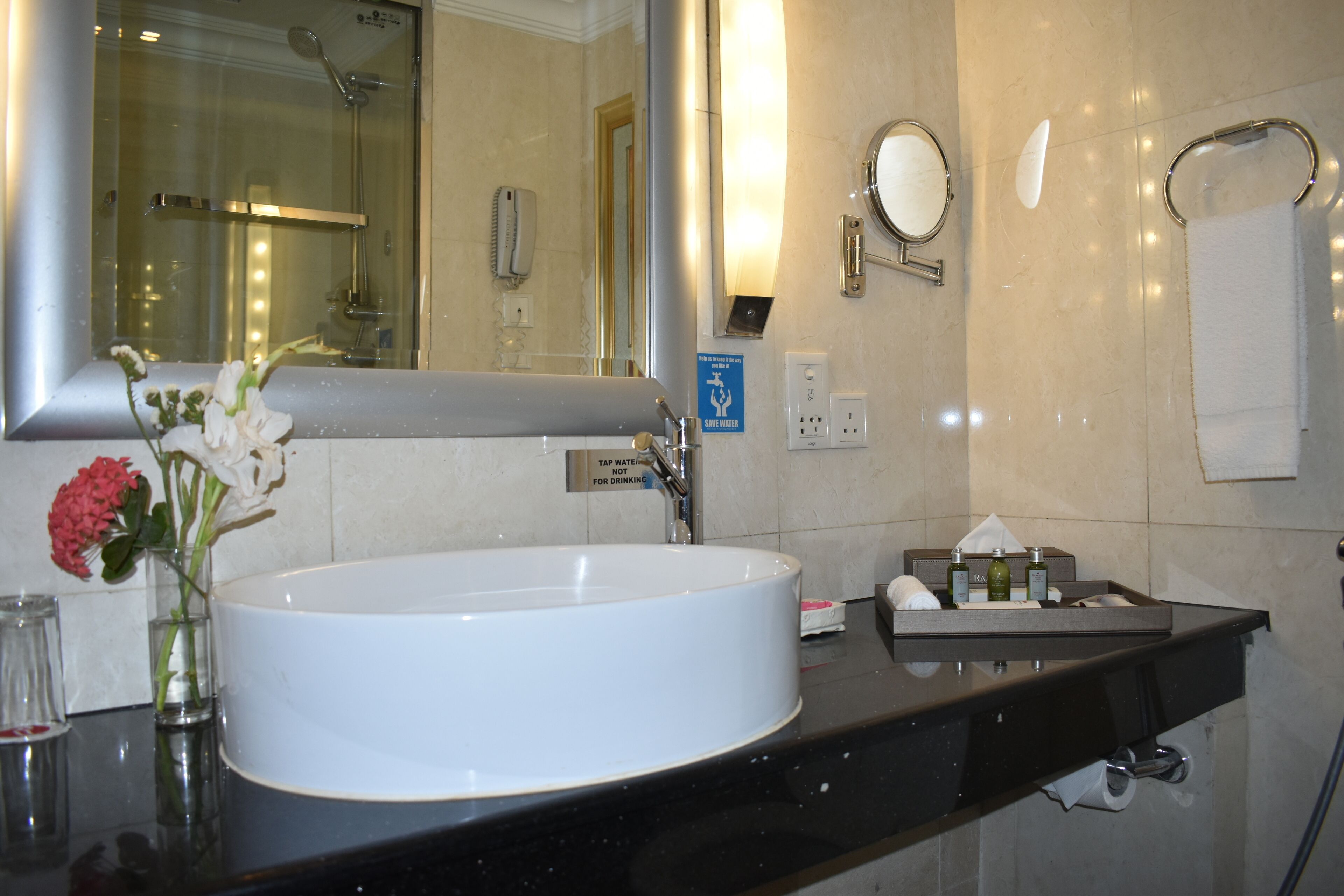 A modern bathroom with a large white sink, a mirror, a glass vase with flowers, and a black countertop.