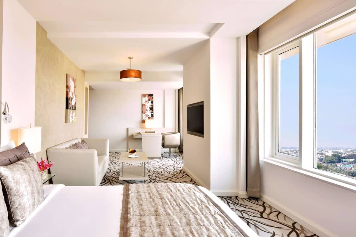 Quarto de hotel com uma cama grande, uma escrivaninha, uma cadeira e uma televisão. Há uma janela com vista para a cidade.