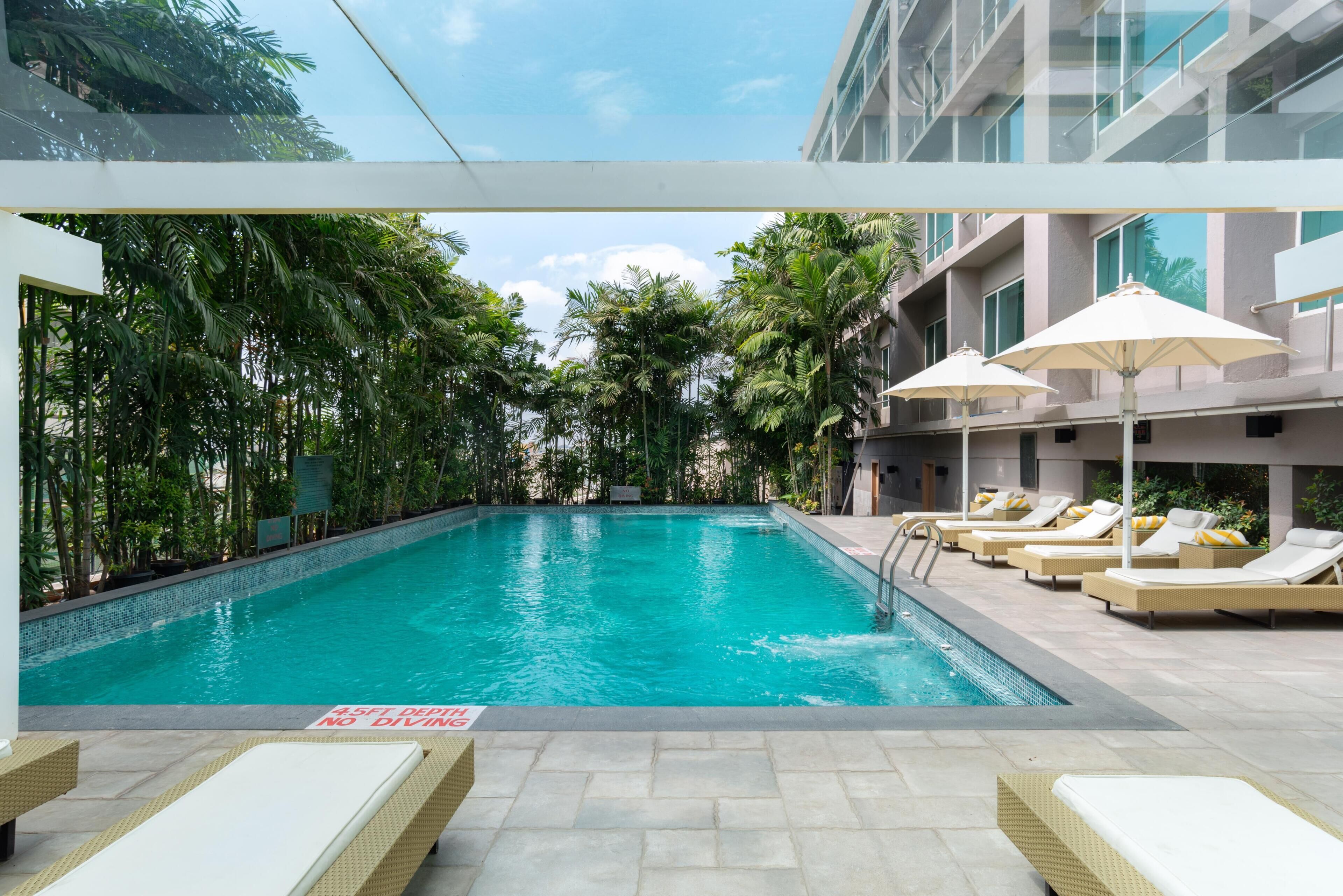 A swimming pool with lounge chairs and umbrellas, surrounded by greenery and modern buildings.