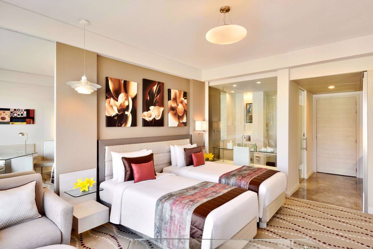 Quarto de hotel moderno com duas camas, um espaço de estar, uma mesa de trabalho e um banheiro visível através de uma porta de vidro.
