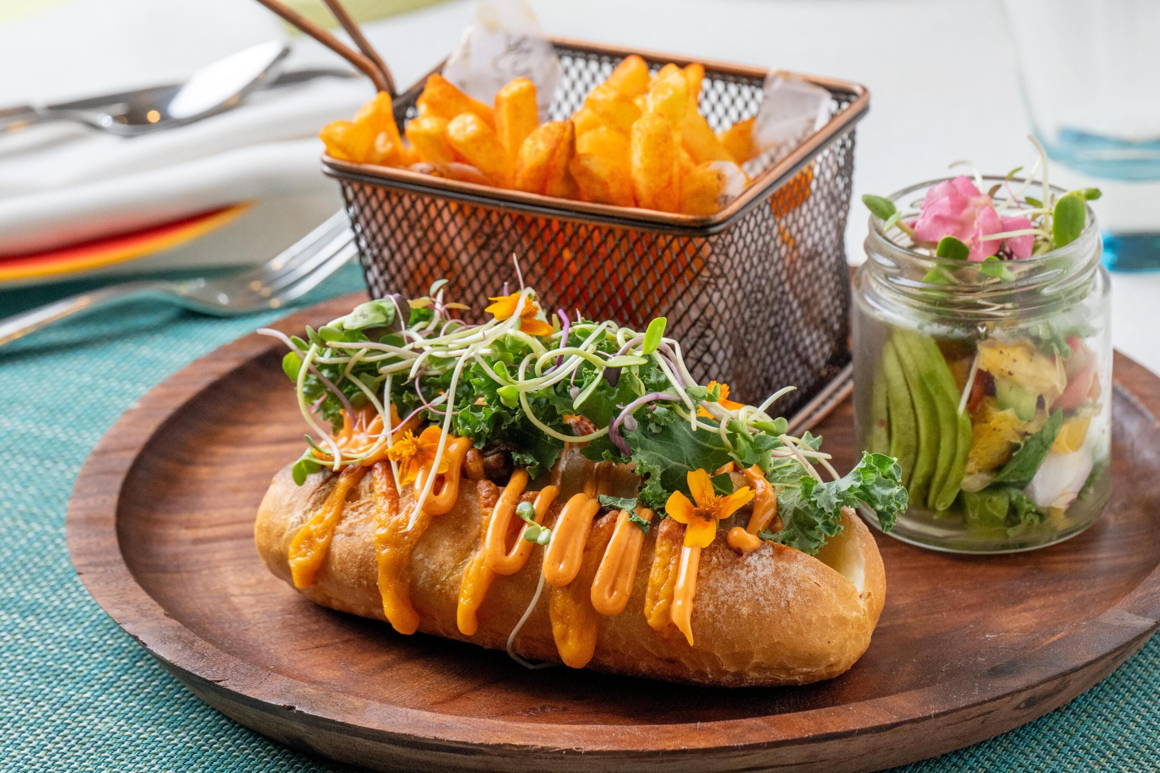 A sandwich with sprouts and sauce on a wooden plate, a basket of fries, and a jar of salad.