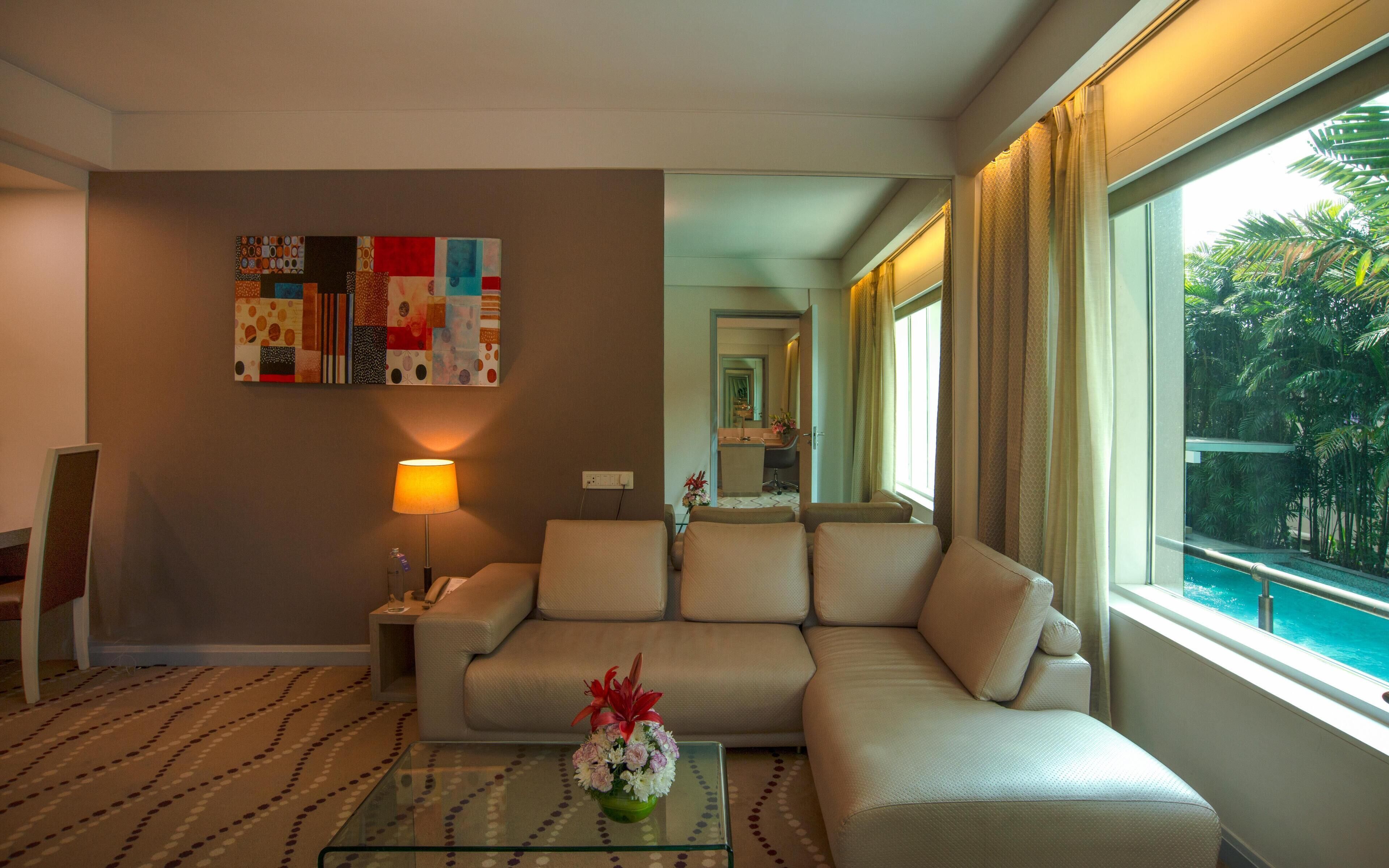 A modern living room with a sofa, a glass coffee table with flowers, a lamp, and a large window with a view of a pool and greenery.