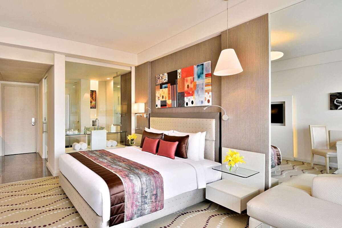 Quarto de hotel moderno com uma cama grande, mesa de cabeceira com abajur, televisão na parede e espelho sobre a cama.