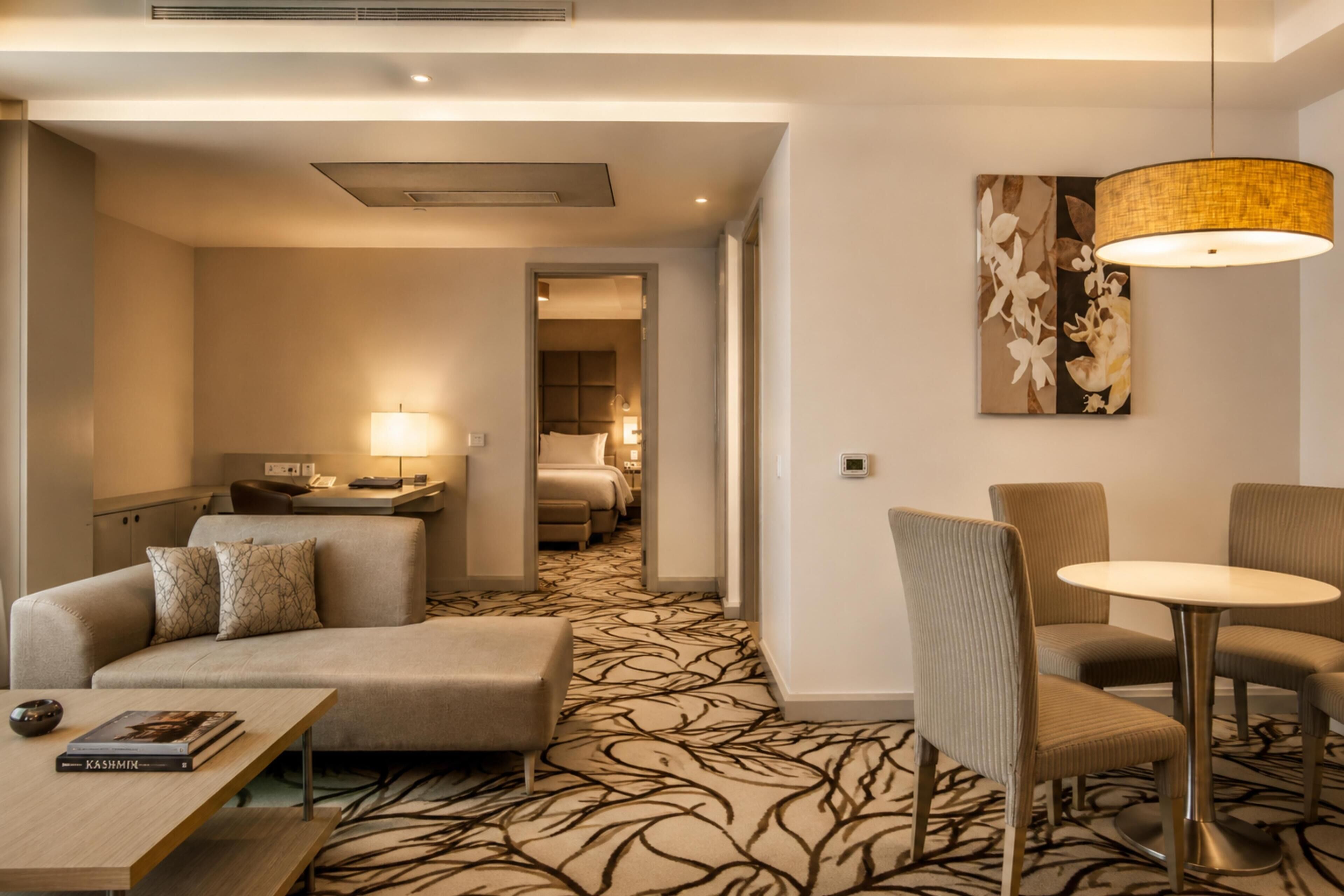 A hotel room with a sofa, a small table, a dining area with chairs, a bed, and a hallway leading to another room.