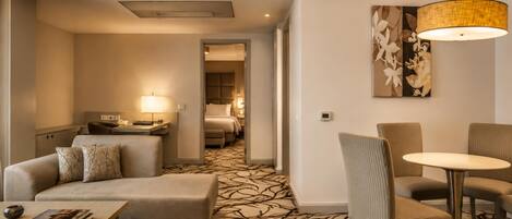 A hotel room with a sofa, a small table, a dining area with chairs, a bed, and a hallway leading to another room.