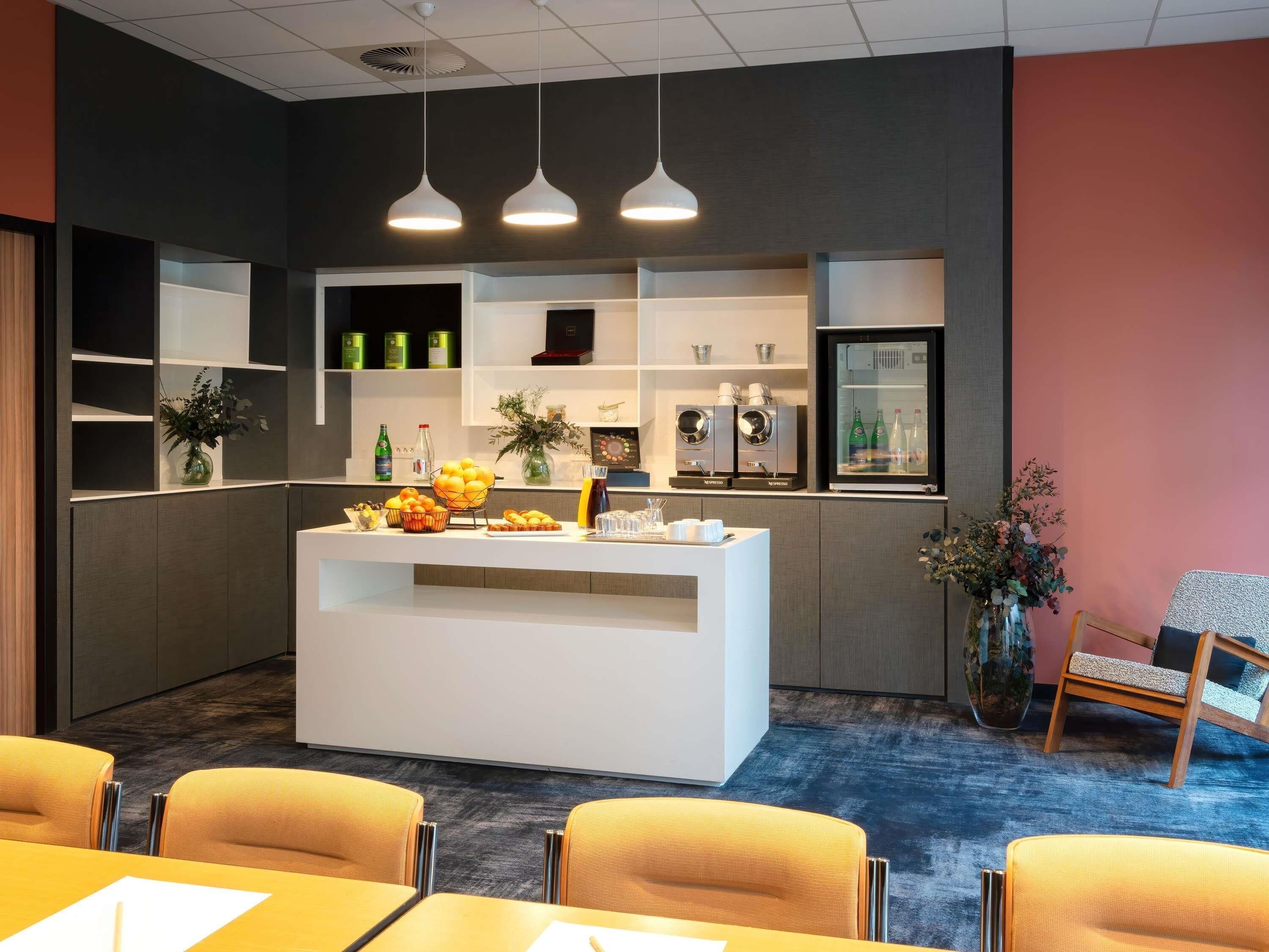 A modern reception area with a white counter, three pendant lights, and a refrigerator. There are yellow chairs and a table with a white placemat.