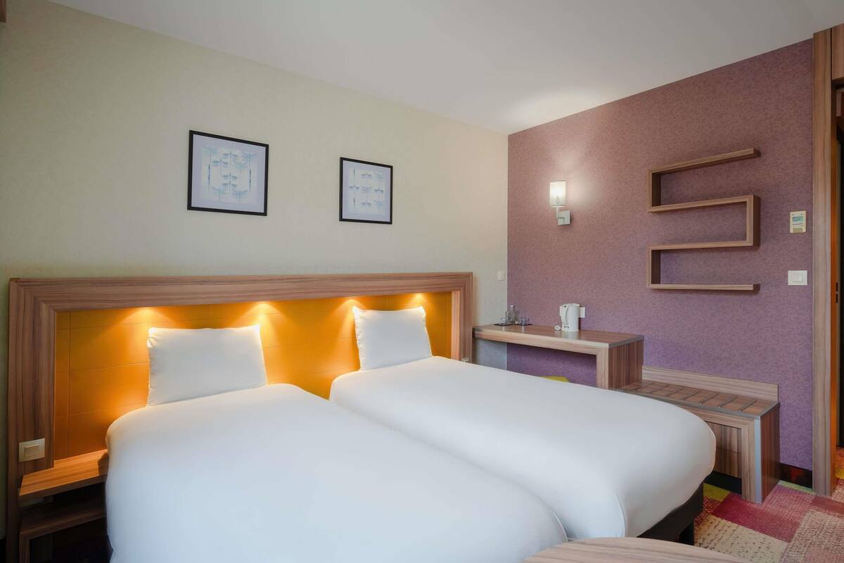 A hotel room with two beds, a wooden headboard, a nightstand with a lamp, and a wall-mounted shelf.