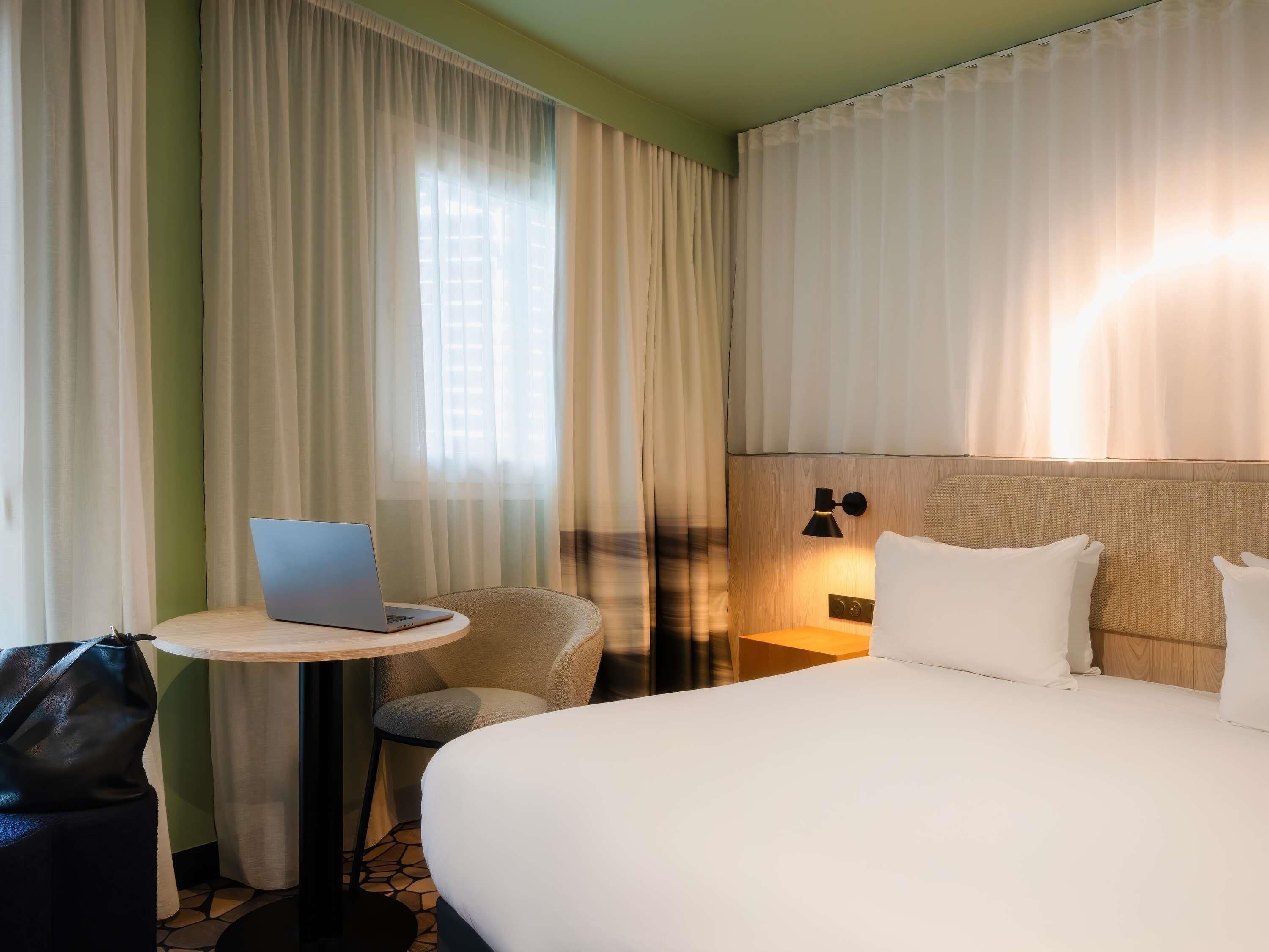Habitación de hotel con cama, escritorio con computadora portátil, silla y mesita de noche con lámpara.