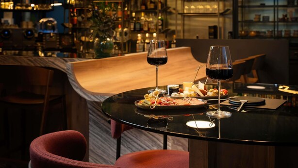 A bar area with a round table set for two, two glasses of red wine, and a variety of snacks and cheeses. - Hotel Mercure Libourne Saint-Emilion (Libourne)