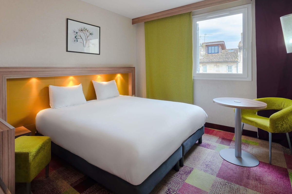 A hotel room with a large bed, a small round table, a green chair, and a window with a view of buildings.