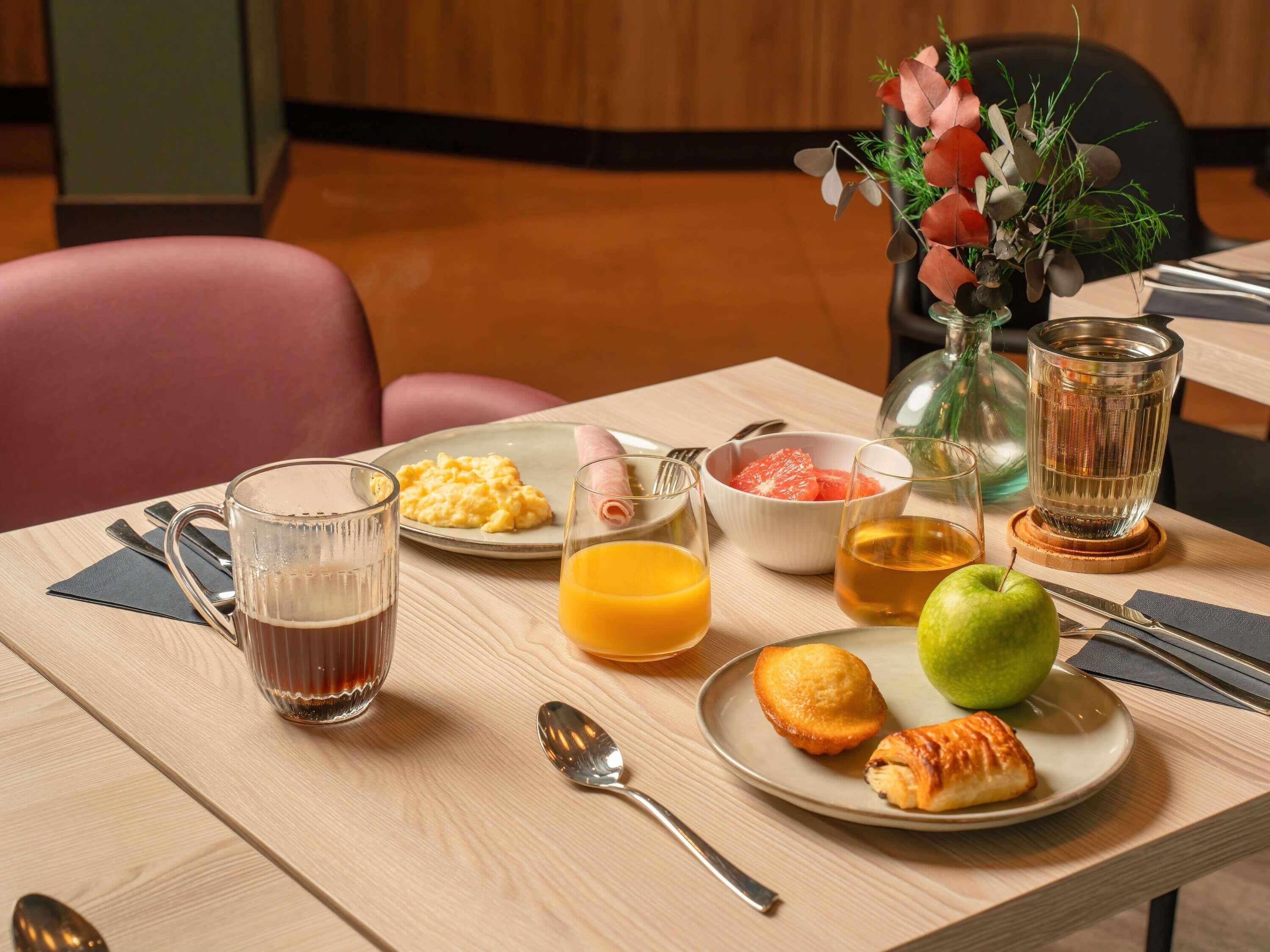 A breakfast table with scrambled eggs, a bowl of fruit, a glass of orange juice, a green apple, a pastry, and a glass of tea.