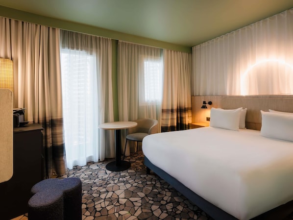 A hotel room with a large bed, a small round table, a chair, and a desk with a lamp.