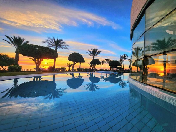 Una piscina con vista al atardecer, rodeada de palmeras y un edificio con ventanales de vidrio reflectante.