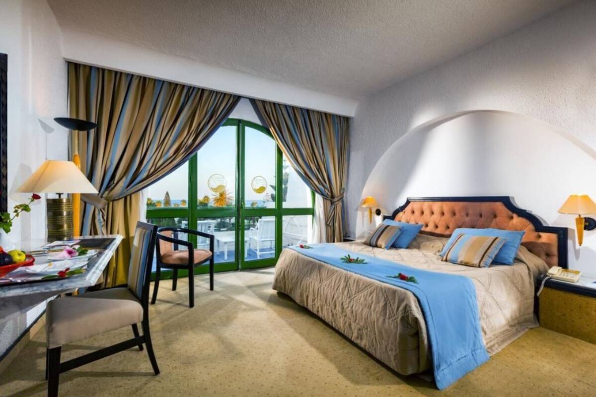 Une chambre d’hôtel avec un lit, un bureau, une chaise et une vue sur l’extérieur grâce à de grandes fenêtres.