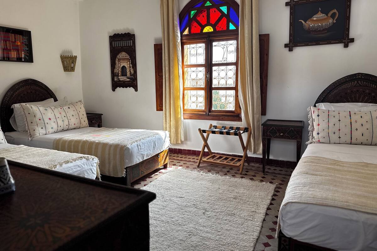 A traditional room with two beds, a small table, a decorative window, and a wall-mounted shelf.