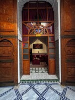 Uma porta de madeira ornamentada com esculturas elaboradas, que dá acesso a um quarto com chão de azulejos estampados e cama com roupa de cama vermelha.