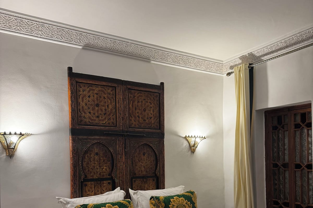 A bedroom with a large bed, ornate wooden headboard, two wall sconces, a nightstand with a decorative vase, and a patterned carpet.